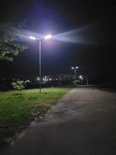 The image depicts an outdoor nighttime scene in the city of Gaba, Nigeria, illuminated primarily by artificial light sources. In the foreground, a tall, slender metallic pole supports a street light assembly featuring two bright, outward-facing LED luminaires, casting significant light and lens flare. To the immediate left of the asphalt pathway, a damp grassy area contains a small, leafy sapling. Overhanging foliage from a larger tree is visible in the upper left quadrant. The asphalt pathway, appearing dark and possibly damp, extends from the foreground towards the background on the right side of the frame. Further down the pathway, additional street lights are visible, casting light onto distant, indistinct structures or buildings in the background. The sky is dark and featureless. No individuals or specific actions are discernible within the frame.