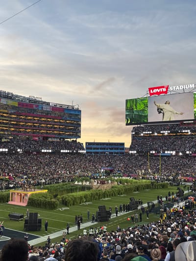 An outdoor stadium is filled with spectators. Levi's Stadium logo and a performer's image are displayed on the large screen. The football field is visible, with some equipment and personnel. The sky is pale blue with some light clouds.