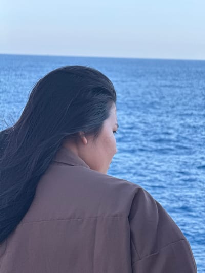 A medium shot photograph captures a woman from the back, her head turned to the right, gazing out at the vast expanse of the sea. She has long, dark hair that cascades over her shoulders and down her back, with some strands caught by a gentle breeze. She is wearing a brown collared shirt. The sea, a deep blue, stretches to the horizon, meeting a pale blue sky with no visible landmass. The overall impression is one of calm contemplation, a moment of quiet reflection against the backdrop of nature's grandeur. The location is the city of Dong Son, Vietnam.