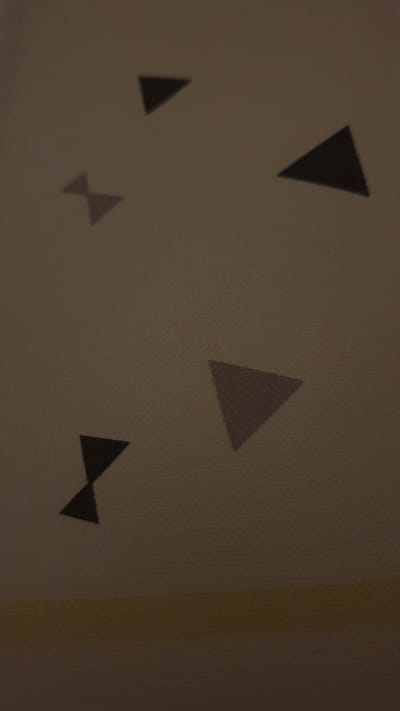 The image shows a patterned surface, possibly a wall or fabric, with several dark geometric shapes resembling triangles and a bow-tie shape. These shapes are scattered across the surface at various orientations and sizes. The lighting in the image is dim, casting shadows and making the details of the pattern less distinct. The surface itself appears to have a subtle texture, possibly a woven material or a textured paint. There is a horizontal band of lighter color, likely a transition to another surface or a painted stripe, at the bottom of the image. The scene does not contain any people, actions, or interactions. There is no information within the image that suggests a specific location in Vrchotovy Janovice, Czechia, or any other geographical context.