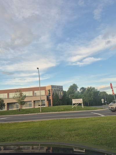 The image captures a view of a modern, two-story brick building with large windows, identified by the "Integrated Research" logo on its facade, located in Dollard-des-Ormeaux, Canada. A street light stands in front of the building. Adjacent to the building, a white sign with text in French and English reads "À Louer / For Lease" and provides a phone number (514) 693-9655. The building is situated on a well-maintained property with a grassy lawn and mature trees. Across a paved road, more green grass is visible, and the reflection of the building can be seen on the shiny surface of what appears to be a car's dashboard or hood in the foreground. A white SUV is parked on the right side of the road, near a stop sign. The sky above is a mixture of blue with scattered white and gray clouds. The overall impression is a commercial or office space in a suburban setting.