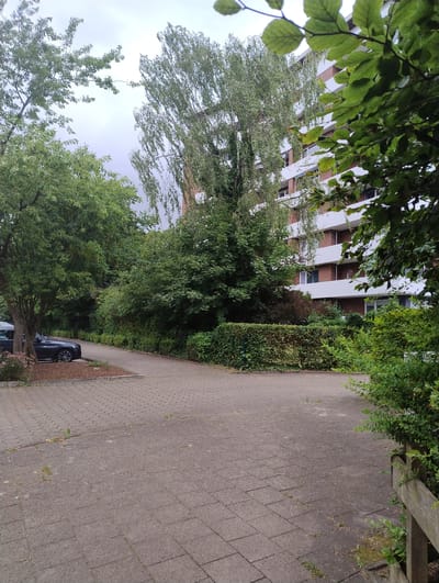 The image depicts a paved area with a parked car on the left, adjacent to a tree. In the background, there is a tall residential building with balconies, surrounded by lush green trees and hedges. A pathway curves away from the foreground into the greenery. The sky is overcast. The scene is likely in a residential area of Uetersen, Germany, given the architectural style of the building and the presence of trees and paved pathways common in German urban environments. No people are visible in the image.