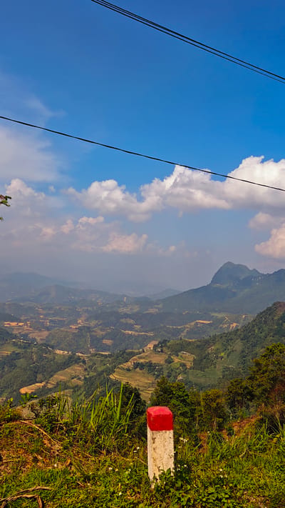 The image depicts a mountainous landscape in Mậu Duệ Commune, Vietnam, under a blue sky with scattered white clouds. Power lines stretch across the top of the frame. Rolling hills covered in lush green vegetation and terraced rice fields extend into the distance. A concrete post with a red painted top stands in the foreground, surrounded by grass and wildflowers.