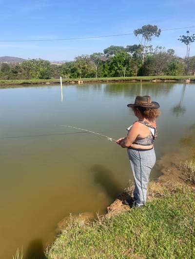 The image depicts an outdoor scene at a pond in Pirenópolis, Brazil, under a clear, cloudless blue sky. An adult female, viewed from a three-quarter rear angle, stands on the earthen bank, engaged in fishing. She wears a brown cowboy hat with a decorative braided band, a sleeveless top with a camouflage-like pattern, and grey plaid trousers with a dark side stripe. Her right hand holds a light-colored fishing rod, with the line extended into the murky, greenish-brown water, producing visible concentric ripples. A single vertical white pole is positioned in the middle distance of the pond. The far bank of the pond and the background are densely vegetated with various green trees. Distant, hazy hills are visible beneath the bright sky, and a thin dark line, consistent with a power line, stretches horizontally across the upper portion of the frame. The immediate foreground consists of green grass transitioning to exposed soil at the water's edge.