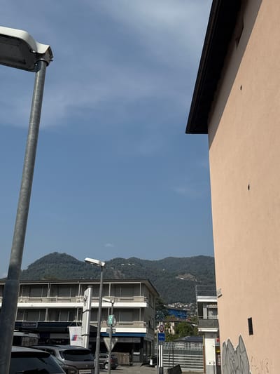 The image captures a view looking up towards a clear blue sky with some wispy clouds. In the foreground, a tall, silver lamppost stands on the left. To the right, the side of a pale pink building with a dark roofline is visible. The building has a rough texture and a small, dark circular vent. Further in the distance, a multi-story building with rows of windows is seen, and behind that, a forested mountain range fills the horizon. Signs for "STAR CAR" and "BMW Premium Selection" are partially visible on the lower levels of the distant building. The scene appears to be in an urban or semi-urban environment, possibly a street or parking area, given the presence of vehicles and commercial signage. The town of Balerna, Switzerland, is indicated by a road sign.