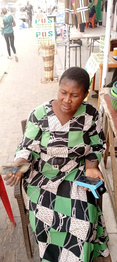 An adult female is seated outdoors on a brown woven chair on a paved street in Kwachiri, Nigeria. She wears a long-sleeved dress with a distinctive green, black, and off-white checkered pattern featuring chains and stylized 'F' shapes. Her short dark hair is visible, and her facial expression suggests concentration, with her mouth slightly open. In her left hand, she holds a stack of folded paper currency. Her right hand grips a light blue and black Point of Sale (POS) terminal.
The background reveals a bustling street scene. To the immediate left of the subject, part of a red chair is visible. Further back, a "POS TRANSFER WITHDRAW DEPOSIT" sign, displaying green and red text, stands prominently on a tall, cylindrical woven wicker base. Beyond the sign, two individuals are visible walking: an adult female in a dark green top and black leggings walking away, and an adult male in a dark t-shirt walking further into the background.
To the right of the subject, a wooden table-like structure and a small black stool are visible. A green and black bowl rests on the table. Hanging textiles and a "MR POOL" barber shop sign are discernible further back on the right. The ground is paved with interlocking grey blocks. The scene is brightly lit by daylight.