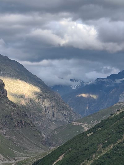 The image captures a dramatic mountain landscape in Gramphu, India, characterized by rugged terrain and a cloudy sky. The foreground is dominated by steep, rocky slopes covered in patches of green vegetation. A prominent band of sunlight illuminates a section of the mountainside on the left, highlighting its texture and creating a stark contrast with the surrounding shadows. In the middle ground, a valley carved by erosion descends, with a winding road visible on its slopes. Further in the distance, layers of mountains recede into the haze, some peaks adorned with snow and shrouded in clouds. The sky above is a canvas of dark, turbulent clouds interspersed with patches of brighter sky, suggesting a dynamic weather system. No people, animals, or specific man-made structures, apart from the road, are discernible. The overall atmosphere is one of wild, untamed natural beauty.