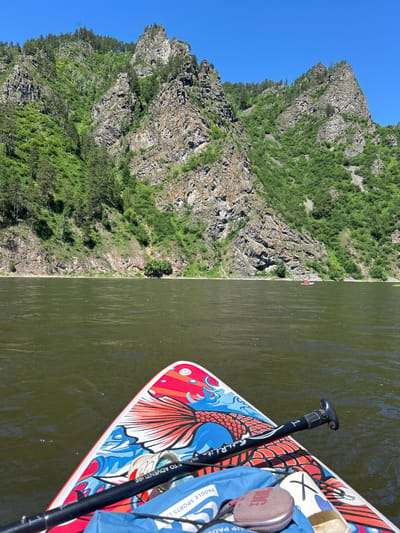 The image shows a paddleboard's nose and paddle on a river with a steep, rocky mountainside covered in green foliage in the background under a clear blue sky. The paddleboard is decorated with a colorful design featuring a fish and waves. A blue bag and a round, mesh-covered object are also visible on the paddleboard. In the distance, on the river, another person is visible on a small red raft. The environment suggests a natural, outdoor setting, possibly a recreational area. The location is identified as Ust-Mana, Russia.