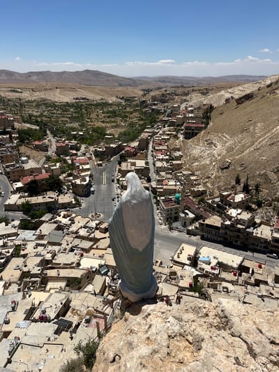 The image is an upward, angled shot from the statue of the Virgin Mary on a cliff overlooking the city of Ma'loula, Syria. The statue's head and draped body are visible in the lower-right, composed of white and blue. The landscape includes a mix of arid, mountainous terrain in the background and dense, light-colored residential buildings, with flat roofs, and narrow streets in the city. Patches of green vegetation are interspersed among the buildings. Several roads are apparent, carrying a light stream of traffic. The sky is clear blue, with a light band of haze.
