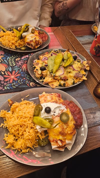 The scene depicts a dining experience in a restaurant located in Aschaffenburg, Germany. Three plates of Mexican-style food are presented on a dark-stained, multi-planked wooden table.
In the foreground, a grey plate holds a cheese-covered enchilada or burrito, topped with a dollop of white cream, a single black olive, several slices of green jalapeño peppers, and one whole green pickled pepper. To its left is a mound of orange-colored rice, and to its right, a portion of red salsa.
Centrally positioned, a second grey plate contains a large serving of nachos. These tortilla chips are covered with melted cheese, chopped red onions, black olives, green zucchini pieces, and a generous layer of green guacamole, garnished with two whole green pickled peppers.
Partially visible in the background, a third grey plate presents a dish identical to the foreground plate.
Two individuals are partially visible in the upper portion of the image. The person on the left wears a white long-sleeved top. The person on the right wears a light beige knitted sweater, with a dark tattoo visible on their right forearm.
The table features a rectangular fabric placemat with a vibrant floral pattern in red, orange, green, and blue. On the right side of the table, a red cylindrical decorative item with an illustration is partially visible, alongside an amber-colored, textured glass candle holder emitting a soft glow. A silver utensil handle is also partially visible in the upper right. The environment is indoors, characterized by warm, artificial lighting.