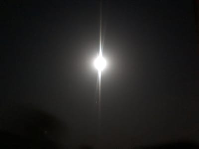 The image depicts a bright, radiant light source, likely the moon, shining intensely against a dark night sky. The light emits a vertical beam or flare, creating a dramatic effect. The surrounding area is shrouded in darkness, with only faint, shadowy shapes visible at the bottom of the frame, possibly indicating foliage or structures. The overall scene evokes a sense of nighttime mystery.