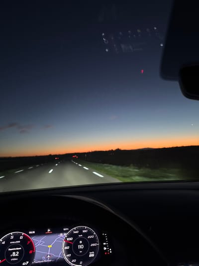 The image presents a first-person view from inside a moving vehicle on a two-lane asphalt road during either dawn or dusk, specifically identified as the D51 road within the Kaysersberg-Vignoble commune, France. The sky exhibits a gradient from dark blue overhead to a distinct orange and red at the horizon, where distant dark landforms are silhouetted. A faint crescent moon is discernible in the upper right quadrant of the sky.
The road ahead is illuminated by the vehicle's headlights, revealing solid white edge lines and dashed white center lines, and extending into the distance where it appears to curve gently left. Green verges are visible immediately adjacent to the road. Multiple red taillights from other vehicles are visible further along the road.
The lower portion of the frame is occupied by the vehicle's digital instrument cluster. It displays the current time as 06:36 and an ambient temperature of 6.0°C. The speedometer indicates the vehicle's speed at 110 km/h, which is above a visible speed limit of 100 km/h. Fuel consumption is shown as 8.2 l/100km. A navigation map on the central display highlights the vehicle's position on the D51 road and labels a point of interest as 'Tourisme Bunner'. Reflections, including a faint red triangular icon and other circular artifacts, are present on the windshield. No individuals are visible within the frame.