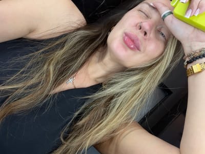 The image shows a close-up, selfie-style shot of a woman lying down and looking towards the camera. She has her eyes partially closed and her lips are pursed, with lip gloss on. She is wearing a black top, a cross necklace, a nose ring, and multiple bracelets on her wrist, including one with beads and a gold watch. She is also wearing several rings on her fingers, one of which appears to be a stack of green and white bands. Her hair is long and appears to be blonde with lighter streaks. She is holding a bright yellow object, possibly a phone case, near her forehead. The background is dark and out of focus, with some reflective surfaces visible. There is no information about the location within Укромновское сельское поселение, Ukraine, or any specific details about the environment.