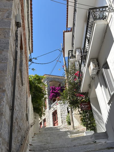 The image depicts a narrow, ascending stone staircase in Skiathos, Greece, flanked by traditional whitewashed buildings. The left side of the staircase is bordered by a rough, grey stone wall with electrical conduits running vertically. On the right, a white building features multiple balconies with ornate black railings and air conditioning units mounted on its facade. Blooming pink and purple bougainvillea vines cascade down from the balconies and an upper terrace, adding vibrant color to the scene. A wooden sign for "Pension Argo" is visible on the right, attached to a wooden pole amidst greenery. The sky above is a clear, bright blue. The scene is illuminated by strong sunlight, casting sharp shadows. There are no people visible in the image.