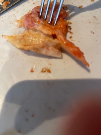 A close-up, overhead shot shows a fork digging into a partially eaten chicken wing on a white plate. The chicken wing appears to be coated in a red sauce, and some crispy skin is visible. There are small crumbs and red speckles scattered across the plate, suggesting that other food was present. A shadow of the fork is cast on the plate to the left, and a larger, blurry shadow, possibly from a hand or an object, occupies the right side of the image. The lighting suggests it is daytime. The overall scene is casual, likely a meal being consumed. No visible text is present in the image.