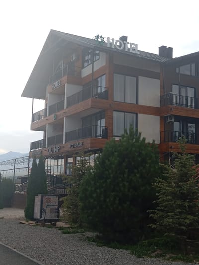 The image shows a multi-story hotel with a wood-paneled facade and balconies. The word "HOTEL" is displayed in large white letters at the top of the building. Below, the words "RESTAURANT OLIMP РЕСТОРАН" are visible on a lower level. The hotel is situated in a green, possibly mountainous, area, with evergreen trees in the foreground. A wheeled cart with a tiled surface is parked on a gravel path in front of the building. String lights are visible in the background. The sky is overcast.