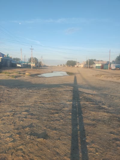 The image depicts a dusty, unpaved road in the city of Aral, Kazakhstan. The road is lined with low-rise buildings, some of which appear to be houses with colorful fences. Power lines crisscross the sky above, supported by wooden poles. A small puddle of water reflects the clear blue sky, which is dotted with a few wispy clouds and the moon. The ground is covered in sand and gravel, with tire tracks visible. A long shadow of a person stretches across the road, indicating the presence of the photographer. The overall scene suggests a rural or semi-urban environment, characteristic of many towns in Central Asia.