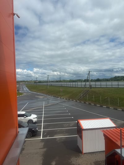 An outdoor, high-angle, full shot shows a parking lot with two cars parked in it next to an orange building. The parking lot is wet and has white painted lines dividing the parking spaces. The cars are a white SUV and a black sedan. There are two small orange and white buildings in the foreground. In the background, there is a metal fence with green grass behind it, and further back, trees and hills. The sky is cloudy. A security camera is mounted on the orange building in the upper left corner. The scene appears to be in the city of Sof'ino, Russia.