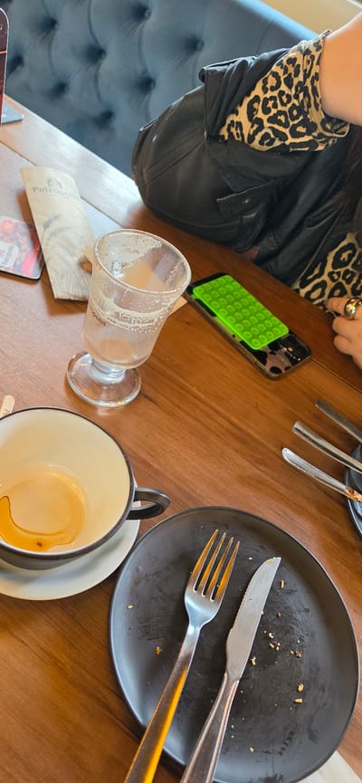 The image shows a table at a restaurant in Nova Petrópolis, Brazil. An individual wearing a black leather jacket and a leopard-print top sits at the table. A partially consumed latte is in a glass, and an empty coffee cup and saucer are also present. A black plate contains leftover food and utensils. A cell phone with a green, textured case is on the table, along with a restaurant menu.