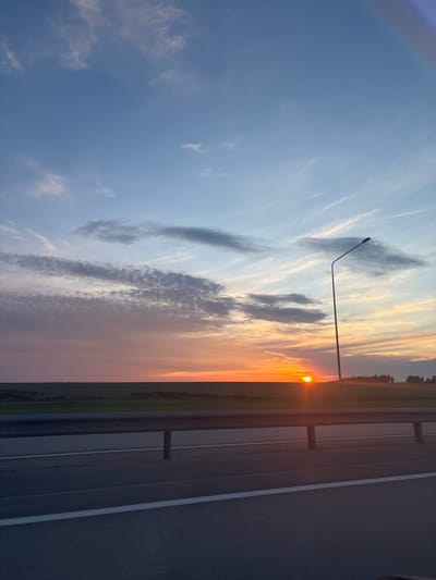 The image captures a scenic sunset over a rural landscape in Новые Карамалы, Russia. The setting appears to be alongside a road or highway, indicated by the asphalt surface and guardrail on the right side of the frame. A solitary lamppost stands tall on the left, silhouetted against the vibrant sky. The sky itself is a canvas of colors, transitioning from a soft blue at the top to warm oranges and yellows near the horizon, where the sun is setting. Wispy clouds, painted with streaks of light, add depth and texture to the celestial display. A dark line of trees marks the distant horizon, separating the sky from the fields below. The overall mood is serene and peaceful, evoking the beauty of nature at dusk. No people or animals are visible in the image, and there is no visible text.