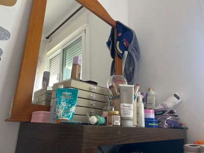 The image depicts a cluttered dresser top with a mirror above it, suggesting a vanity area. On the dresser, various cosmetic and personal care items are arranged. Prominent objects include tubes of sunscreen, makeup brushes in a container, bottles of lotions and moisturizers, a makeup organizer with drawers, and a small jar. A blue and red baseball cap with "VSTAR BOGOTA" is hanging on the frame of the mirror. The overall impression is one of a functional, used personal space, likely in a residential setting. The mention of "Pueblo Salinas, Spain" in the prompt implies this scene is situated there, though no specific visual cues in the image directly confirm this location. The lighting suggests it is daytime, possibly with natural light coming from a window visible in the reflection of the mirror.