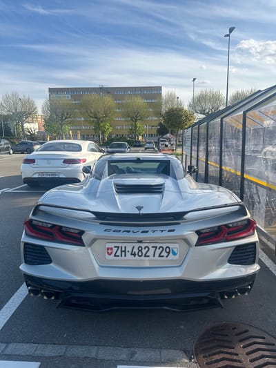 An overhead, slightly angled rear view captures a silver Chevrolet Corvette C8 parked outdoors on a sunny day in what appears to be a parking lot or street in Volketswil, Switzerland. The car is oriented with its rear facing the viewer. The Swiss license plate "ZH-482 729" is clearly visible on the back, featuring the Swiss flag emblem. The "CORVETTE" emblem is emblazoned across the rear bumper. The car has distinctive red taillights and quad exhaust pipes visible on the right side.
Behind the Corvette, a white Mercedes-Benz AMG with the Swiss license plate "ZH-596 889" is also parked. Other vehicles are visible further in the background. To the left of the Corvette, a transparent bus stop or bike shelter structure can be seen. On the right side of the Corvette, a round metal manhole cover is set into the asphalt.
In the background, under a bright blue sky with scattered white clouds, a large, light-colored, multi-story building stands. The word "imicasa" is visible on the side of this building. Several trees with light green foliage are interspersed between the parked cars and the building. Streetlights are also visible against the sky. The time of day is daytime, with clear weather. No people are visible in the image.
