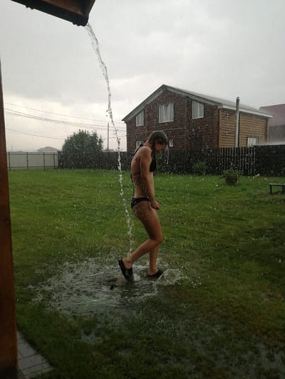 In Yemelyanovo, Russia, a person in a black bikini stands in a grassy yard, receiving a stream of water from an overhead gutter. The water cascades down, creating puddles on the lawn. In the background, a two-story wooden house with visible windows and a fence are situated. The sky is overcast, and raindrops are evident throughout the image, suggesting it is raining. The scene captures a moment of outdoor activity amidst inclement weather.