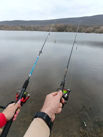 The image depicts two people fishing in a body of water near Орлиновский округ, Ukraine. Each person holds a fishing rod, one with a red and black reel and the other with a green and black reel. Both rods are rigged with fishing line and lures. The background shows a calm lake surrounded by trees and hills under an overcast sky. One person wears a beige sleeve and a black smartwatch, while the other wears a red sleeve and has pink manicured nails. A piece of trash lays in the grass near the edge of the water.