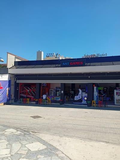 The image shows a street-level view of an arcade and a hotel in Chalkida, Greece, on a clear, sunny day. The arcade, named "SAT GAMES," is a storefront with an open-front design displaying various arcade machines. Red, green, and yellow stools are arranged in front of the arcade, with a few people seated at some of them. One person in a white shirt is sitting at a stool and looking towards the arcade entrance. Inside the arcade, other individuals can be seen interacting with the games. A large blue banner with white text is visible on the left side of the frame. Above the arcade, a sign for "LUCY HOTEL" is prominently displayed, indicating the location. The street in front of the arcade is paved with interlocking bricks and large stone slabs. The overall scene depicts a commercial area with leisure and hospitality businesses.