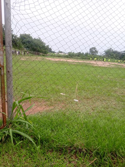 The image shows a fenced-off grass field, likely a sports field, in the city of Ife, Nigeria. In the distance, several people are visible on the field, some wearing yellow shirts, suggesting a sports activity like football. The field is partially covered in grass and partially in dirt. The foreground shows dense green grass and a portion of a metal fence post, indicating the viewer is observing the scene from outside the field. The sky is overcast, with a bright, washed-out appearance. The overall scene conveys an outdoor, casual setting, possibly a training session or a game.