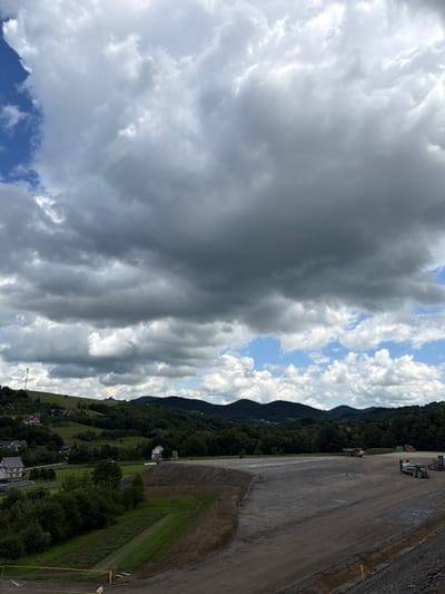 The image depicts a large, open, dirt area that appears to be a construction site or landfill. Several pieces of heavy machinery are present, including a yellow dump truck, a green tractor with a trailer, and a forklift. In the background, rolling green hills are covered with trees, and several houses and buildings are visible scattered across the landscape. A tall transmission tower stands on one of the hills. The sky is partly cloudy with dramatic, dark clouds dominating the upper portion of the frame. The scene is likely located in the rural outskirts of Janowice, Poland, given the presence of the characteristic hilly terrain and vegetation. The overall impression is one of ongoing development or land management in a natural, scenic environment.