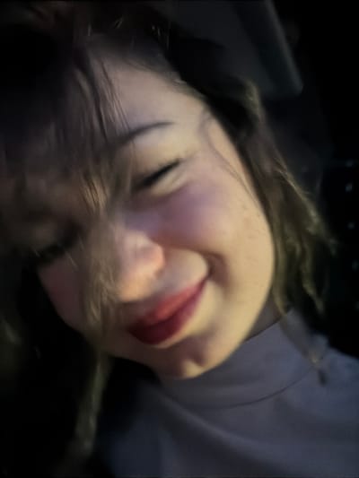 A close-up, slightly blurred shot depicts a person with fair skin, dark hair, and freckles. Their eyes are closed, and they have red lipstick on. They are lying down against a dark surface. The angle is canted, making the person appear sideways. A light grey fabric is visible behind the head. The lighting is dim and unclear, with some dark shadowed areas, resulting in a somewhat obscured image.