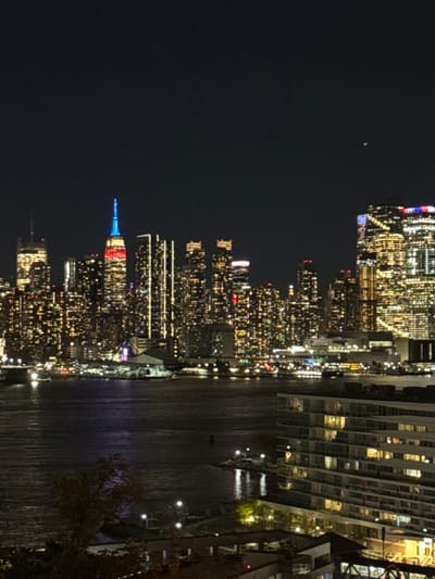 The image displays a nighttime long shot of the Manhattan skyline, viewed across the Hudson River from an elevated perspective in Weehawken, United States. The dark night sky features a single, faint reddish-orange light in the upper right quadrant.
The distant skyline is composed of numerous illuminated skyscrapers. The Empire State Building, centrally positioned to the left, is distinctly lit in red, white, and blue. Other high-rise structures display dense patterns of warm, yellow interior lighting and vertical exterior accent lights, forming a brightly lit urban environment.
The Hudson River occupies the midground, presenting a dark surface with diffuse reflections of city lights, including faint blue and purple tones. Small, distant light sources are discernible on the water.
In the lower right foreground, a multi-tiered, modern building with numerous illuminated windows and balconies is prominent, indicating an active interior. The immediate lower-left foreground includes dark tree foliage and additional ground-level lights and indistinct structures along the Weehawken waterfront. No human figures or specific actions are discernible.