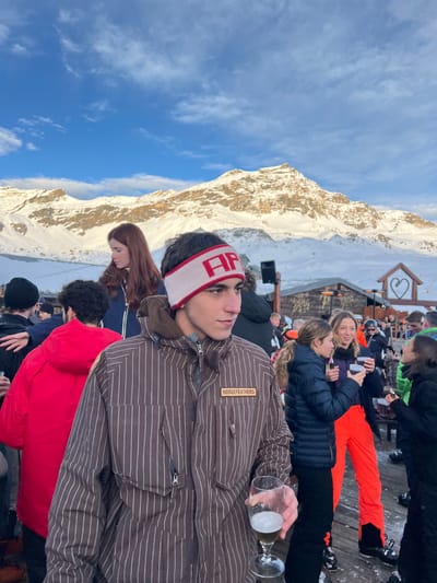 The scene depicts a crowded outdoor après-ski gathering in Valtournenche, Italy, set against a backdrop of snow-covered mountains under a bright blue sky with sparse clouds. In the immediate foreground, slightly off-center, a young male with dark hair wears a white headband featuring red "AP" lettering, a brown and white pinstriped ski jacket with a "HORSEFEATHERS" patch on the left chest, and holds a stemmed glass containing a light-colored beverage. He looks towards the right of the frame.
To his left and slightly behind, another male is visible from the back, wearing a red winter jacket. A female with long red hair, dressed in a dark blue jacket, stands further back, looking to her left. To the right of the main subject, a group of individuals mingles, including two females in dark jackets, one wearing bright orange ski pants, holding dark mugs.
The background features a rustic wooden structure, possibly a hut or bar, with a decorative heart-shaped cutout on its exterior wall. A black speaker box is visible on a stand near the structure. Numerous other individuals, dressed in various winter ski apparel, are present throughout the mid-ground and background, standing on what appears to be a wooden deck or platform. The snow-covered slopes and peaks of the mountainous terrain dominate the distant background under a clear, bright sky.
