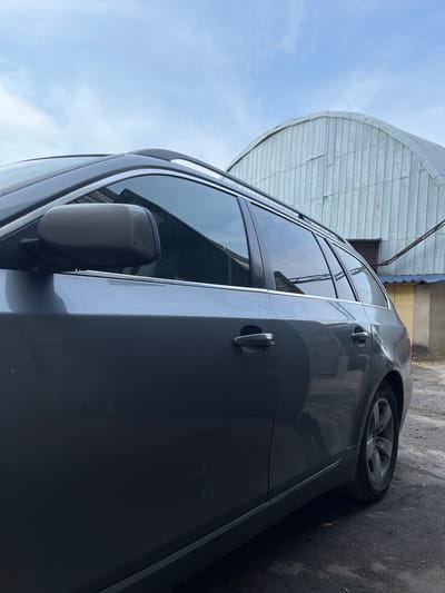 The media file shows the side of a dark gray car parked outdoors. The car is viewed from a low angle, with its side doors and rear wheel visible. The car appears to be a sedan or wagon. The setting is outdoors, with a clear blue sky and some white clouds visible in the upper left quadrant. To the left of the car, a portion of a large, curved, metallic structure is visible, possibly a building or a roof. The ground is asphalt. The image appears to have been taken during the day, likely in the morning or afternoon, given the lighting. There are no people visible in the image. The overall impression is one of an urban or suburban environment. There is no visible text in the image.