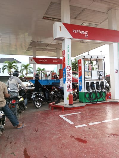The image depicts a gas station with multiple vehicles, including motorcycles and a truck, lined up for refueling. A Pertamax 92 sign is prominently displayed, along with other fuel type labels like BIOSOLAR and DEXLITE. The scene appears to be set in a town or city in Indonesia, possibly Tocina, given the context. Several individuals are visible, some on motorcycles and others attending to the vehicles. The gas station is equipped with fuel pumps, a fire extinguisher, and clear directional markings on the red-colored floor. The environment suggests a typical day at a busy fuel station.