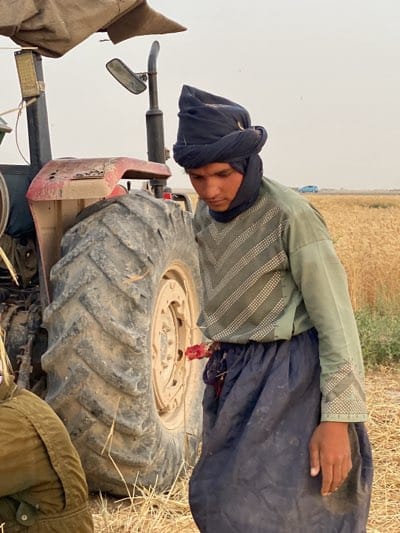 The image shows a man standing next to a tractor in a wheat field, potentially in 'Alaqadari Daman, Afghanistan. He is wearing a dark blue turban, a striped green and white shirt, and a dark blue skirt. He is looking down and appears to be holding something red. The tractor is red and dirty, with a large tire visible. In the background, there is a wheat field and a hazy sky. A light blue car is on the road. The scene suggests an agricultural setting.