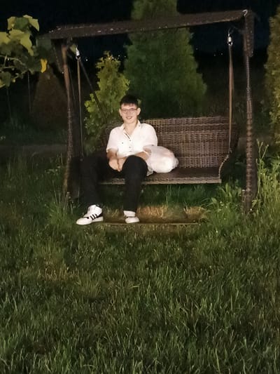 A young man wearing glasses, a white short-sleeved shirt, black pants, and white high-top sneakers with black stripes is sitting on a wicker swing. He is holding a white plastic bag in his lap. The swing is suspended from a dark brown frame and is set in a grassy area with trees in the background. The lighting suggests it is nighttime. This scene is located in Tserovani, Georgia.