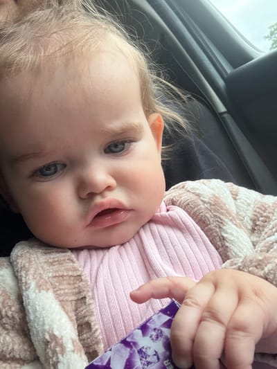 A close-up, eye-level shot shows a baby girl with blonde hair and blue eyes sitting in a car seat. She is wearing a pink ribbed shirt and a fluffy pink and white striped cardigan. Her left hand is holding a small purple object, possibly a candy wrapper. The background shows the interior of a car, including the dark seat and a glimpse of the sky and trees through the window. The image does not provide any information about the location in Braunfels, Germany.