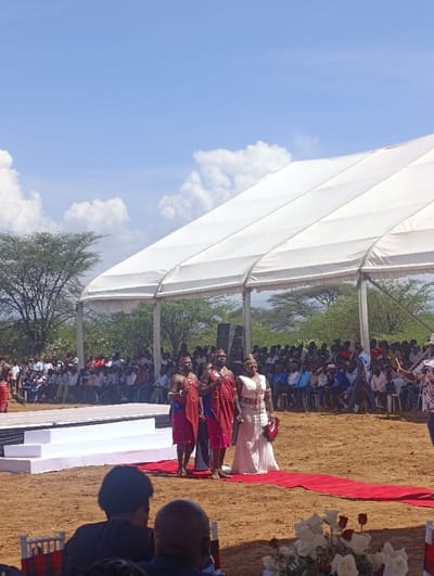 An outdoor ceremonial event is depicted in Marigat, Kenya, under a large white temporary tent. The scene is set on reddish-brown earth. In the mid-ground, a woman in a white gown adorned with beadwork and a crown-like headpiece stands on a red carpet. She holds a dark fan with red accents in her left hand, and her right hand appears to be linked with a man beside her. Flanking her are two men dressed in traditional red shukas, intricate beaded necklaces, and headwear, each holding a staff. To the left of these individuals is a white multi-tiered stage platform. In the background, a large audience is seated on chairs beneath the tent, facing the stage. Sparse green trees line the horizon under a clear blue sky with scattered white clouds. In the immediate foreground, the backs of several attendees' heads are visible, along with a floral arrangement featuring white and red roses on a surface to the right.