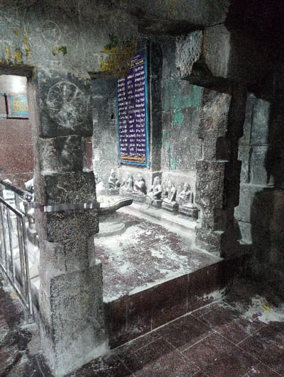 The image displays an interior section of a stone structure, likely a temple, located in Kadaladi, India. The scene is characterized by heavy, dark grey stone architecture featuring multiple pillars and walls.

In the mid-ground, a raised platform with a reddish-brown, speckled surface is visible, heavily dusted with a white powdery residue. Centrally on this platform rests a stone Shiva Linga structure, consisting of a cylindrical lingam positioned within a larger yoni base. To the right of the linga, a row of six smaller, grey stone statues depicting seated figures are arranged along the wall on the raised platform. These statues also show traces of the white powdery residue.

Mounted vertically on the dark grey stone wall above the statues is a blue rectangular plaque with white text, likely in a local script such as Tamil. The stone surfaces of the pillars and walls exhibit rough textures, some greenish discoloration, and signs of age.

On the left side of the frame, a metal railing, possibly indicating a staircase or a designated pathway, is partially visible. The foreground features a tiled floor with a reddish-brown hue, also partially covered with the white powdery residue. The lighting in the scene is dim, casting shadows and highlighting the aged appearance of the stone elements. No individuals are present in the frame.
