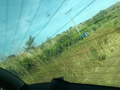 The image, taken from within a vehicle looking through a front or rear window, depicts a hillside environment in Jibilik, Nigeria. The upper portion shows a clear, light-blue sky bisected by multiple diagonal power lines. The middle and lower sections are dominated by a sloping terrain covered in dry, light-brown tall grass and interspersed with greener shrubs. A wooden utility pole, slightly angled, stands on the upper part of the hillside, with electrical wires extending from its top. Below the utility pole, situated on the incline and partially obscured by vegetation, is an object identified as a small, overturned vehicle or utility cart, displaying prominent blue, white, and red color sections. No individuals are visible within the frame. The scene presents a static, outdoor view of a roadside or rural area.