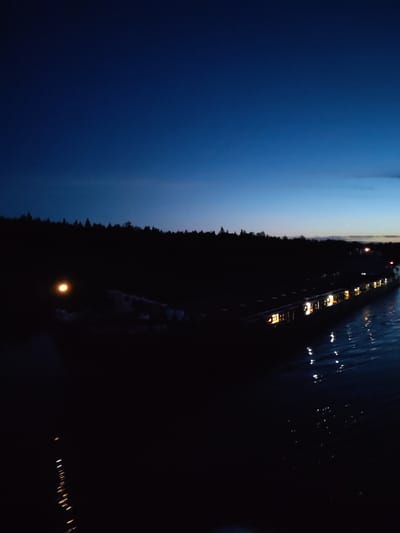 The image depicts a night scene. The sky transitions from a dark blue at the top to a lighter blue/gray near the horizon. A dark, silhouetted treeline stretches across the middle of the image. The foreground is predominantly dark, possibly water, with a line of light reflections suggesting ripples or small waves. A long building or series of buildings with lighted windows runs along the right side of the image, partially obscured by darkness. The specific location in Uzhla, Russia, is not discernible from the image.