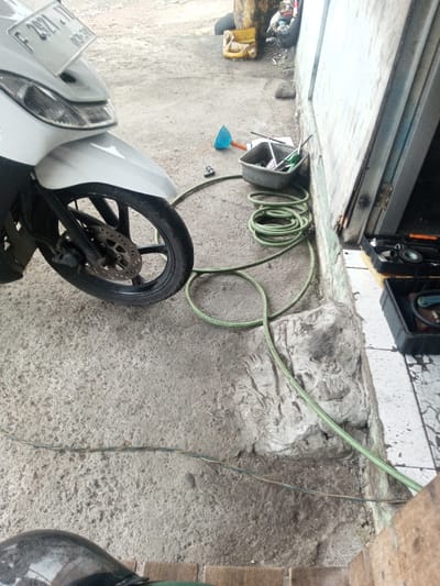 A motorbike parked beside a building features prominently in the shot. Near the building's base, a coiled green hose rests on concrete and extends towards the lower right. Adjacent to the hose are a grey container with tools and a small blue object. A black tool case sits by the building's open doorway. The surrounding area appears to be a workshop or outdoor repair space. A pair of thin electrical wires traverse the ground.