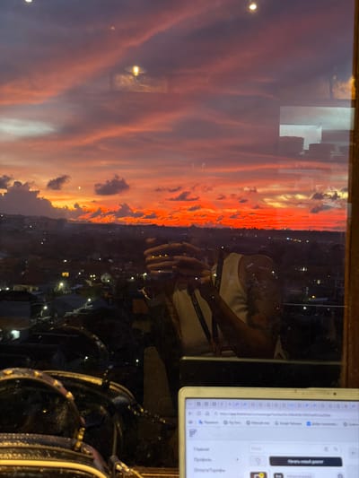 The image captures a panoramic sunset over the city of Benoa, Indonesia, viewed through a window that simultaneously reflects an indoor scene. The sky is illuminated with intense red, orange, and pink hues across numerous clouds, fading to purple at the top, indicative of late dusk. Below the horizon, a sprawling urban landscape of Benoa is visible, dotted with numerous artificial lights from buildings and streets.

Reflected on the window surface, a person's upper torso and arms are discernible. The individual wears a light-colored ribbed tank top, with a visible tattoo on the left arm. Both hands are raised, holding a device, likely a smartphone, with visible light reflections on the fingertips, suggesting the act of photography. A dark strap runs across the person's chest. In the lower right foreground, a white open laptop displays a webpage containing Russian text, including 'Главная', 'Профиль', and 'Начать новый диалог', with a URL indicating 'app.flowdesk.io'. A dark bag with metallic embellishments is visible to the left of the laptop. A reflected interior light fixture appears as a bright, elongated shape near the top center.