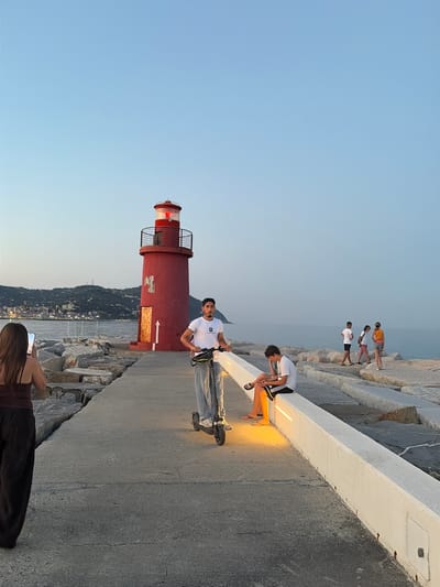 The image shows a group of people on a paved walkway next to a rocky breakwater. In the foreground, a red lighthouse with a black railing and a red light is visible. A man in a white t-shirt and jeans is standing next to the lighthouse, holding a selfie stick. A woman in a dark top is in the lower right corner of the image, taking a photo with her phone.
Further down the walkway, two young men are sitting, one in a white t-shirt and black shorts with Adidas stripes, and another in a white t-shirt and jeans, both illuminated by a warm yellow light. In the background, several other people are walking or standing on the breakwater. The scene is set against a clear blue sky, suggesting it is daytime, likely late afternoon or early evening given the warm lighting. The presence of the lighthouse, breakwater, and the distant town with lights suggests a coastal location. The water is calm. The overall mood appears relaxed and casual, with people enjoying the scenery and taking photos. The location context of Imperia, Italy, further supports the coastal setting.