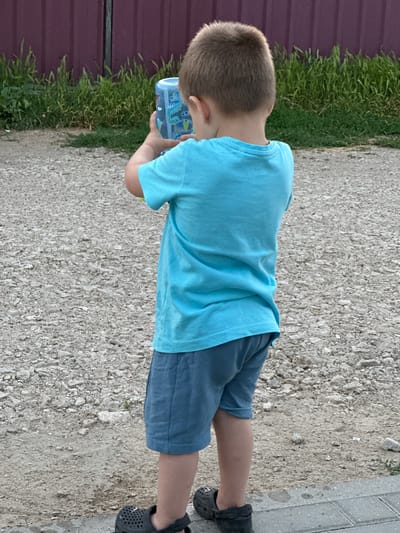 A young boy is standing on a gravelly surface, wearing a light blue t-shirt and blue shorts. He is holding a clear plastic sippy cup with a blue lid, decorated with cartoon dinosaurs and the word "UP". His hair is short and brown. To his left, a dark red fence runs along the edge of a patch of green grass. The ground is mostly gravel and dirt, with some evidence of paving stones to the right. The lighting suggests it is daytime, and the overall scene appears to be outdoors in a casual setting, possibly a backyard or a path.