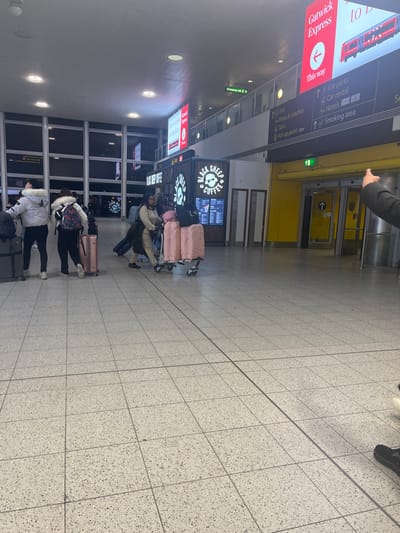 The image depicts an interior view of an airport terminal, likely Gatwick Airport in Horley, United Kingdom, given the prominent "Gatwick Express" signage. The scene appears to be in the evening or night, with artificial lighting illuminating the area and dark conditions visible through large windows in the background.
Several individuals are present, predominantly seen from behind or from a distance, most of whom are accompanied by various pieces of luggage, including wheeled suitcases and backpacks. They appear to be in transit, possibly waiting or navigating through the terminal.
Notable objects include a "BLACK SHEEP COFFEE" shop with a counter and menu boards, around which some people are gathered. Large digital display boards provide directional information, clearly visible text includes "Gatwick Express" with an arrow pointing "This way," and other services such as "Buses & coaches," "Car parks," "Car rental," "Hotels," "Pick-up point uber," and "Smoking area." The floor is covered with light-colored speckled tiles, marked with grid lines. Yellow wall panels and doors are visible in the upper background, with an "exit" sign above a doorway. The overall impression is one of a busy, functional travel hub.
