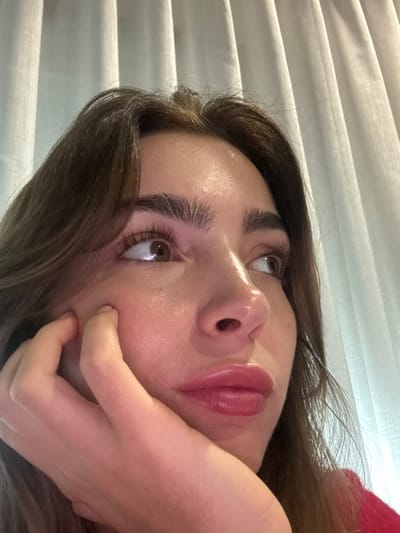A woman's face dominates the frame. She has brown hair, arched eyebrows, and a fair complexion. Her eyes look upward, reflecting light. Full, pink lips are prominent. Her hand is positioned against her cheek and jawline, with fingers slightly curved. A textured, horizontal pattern, likely from blinds, appears in the background. The image is captured at a low angle, looking up towards the woman's face.