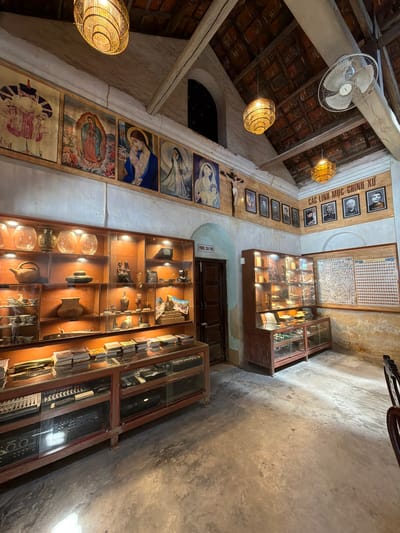 The image depicts the interior of a building that appears to be a museum or a religious exhibition space in Hội Tín, Vietnam. The room has a rustic aesthetic with exposed wooden beams and a tiled roof. Various religious paintings of what seem to be the Virgin Mary and other figures adorn the upper walls.

In the foreground, there are several display cases filled with artifacts. One large display case runs along the left wall, showcasing a collection of what appear to be vases, pottery, and possibly religious items. Below this, another long display case contains what looks like electronic equipment, possibly audio mixers or synthesizers, arranged neatly.

To the right, a taller display cabinet is filled with smaller objects, including what seem to be figurines and other decorative items. Adjacent to this cabinet, a large framed display board features a grid of numerous small photographs or stamps.

The space is illuminated by warm, artificial lighting from hanging lamps and spotlights within the display cases, casting shadows on the concrete floor. A wooden door is visible on the right side of the room, and a ceiling fan is mounted near the roof. The overall atmosphere is one of historical preservation and cultural display.